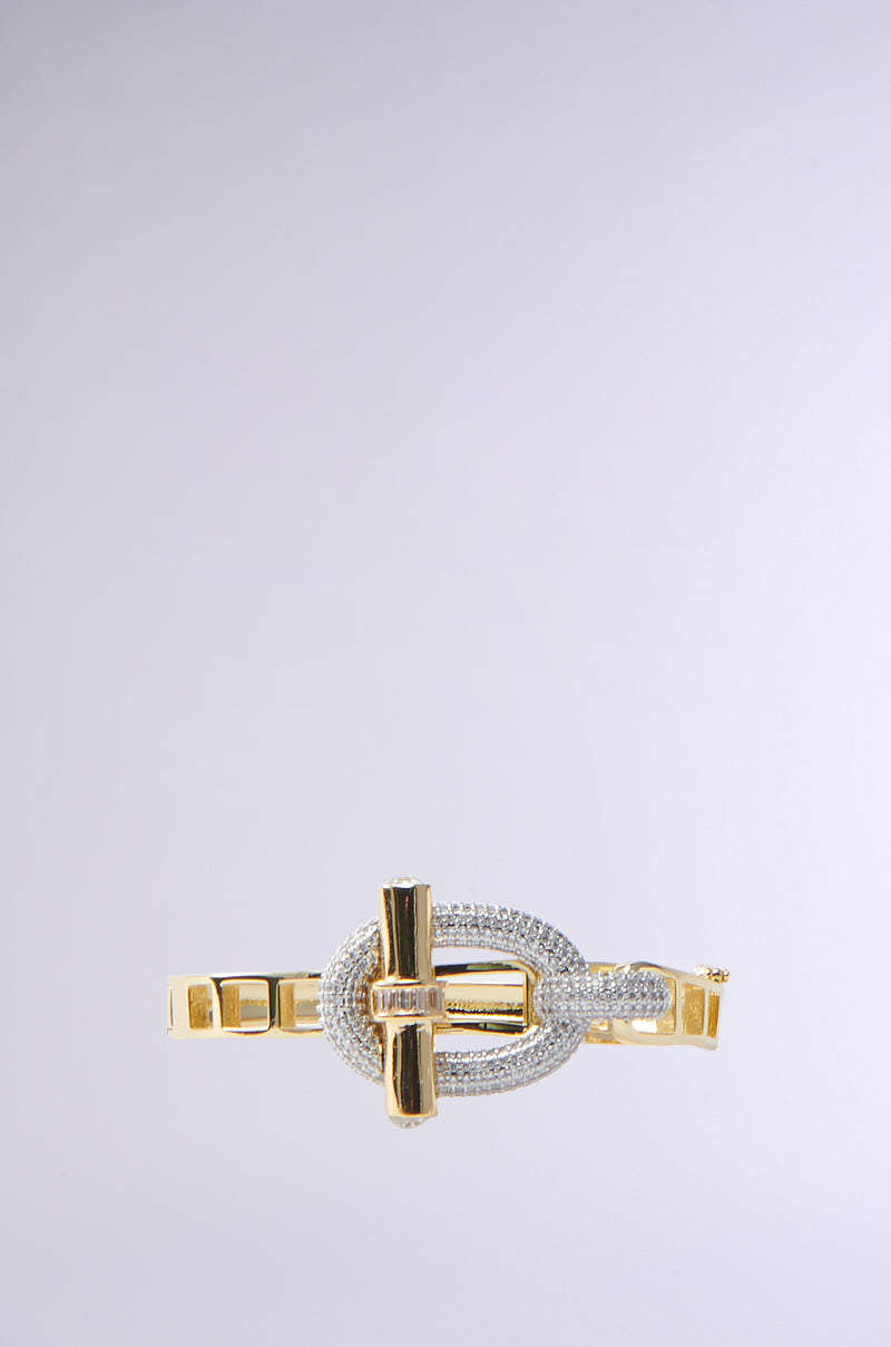 The IYKYK Bracelet by AKIRA Label is a gold treasure featuring a unique loop design, embellished with diamonds and rhinestones. Its mix of smooth and textured finishes creates an elegant, luxurious look against a neutral background.