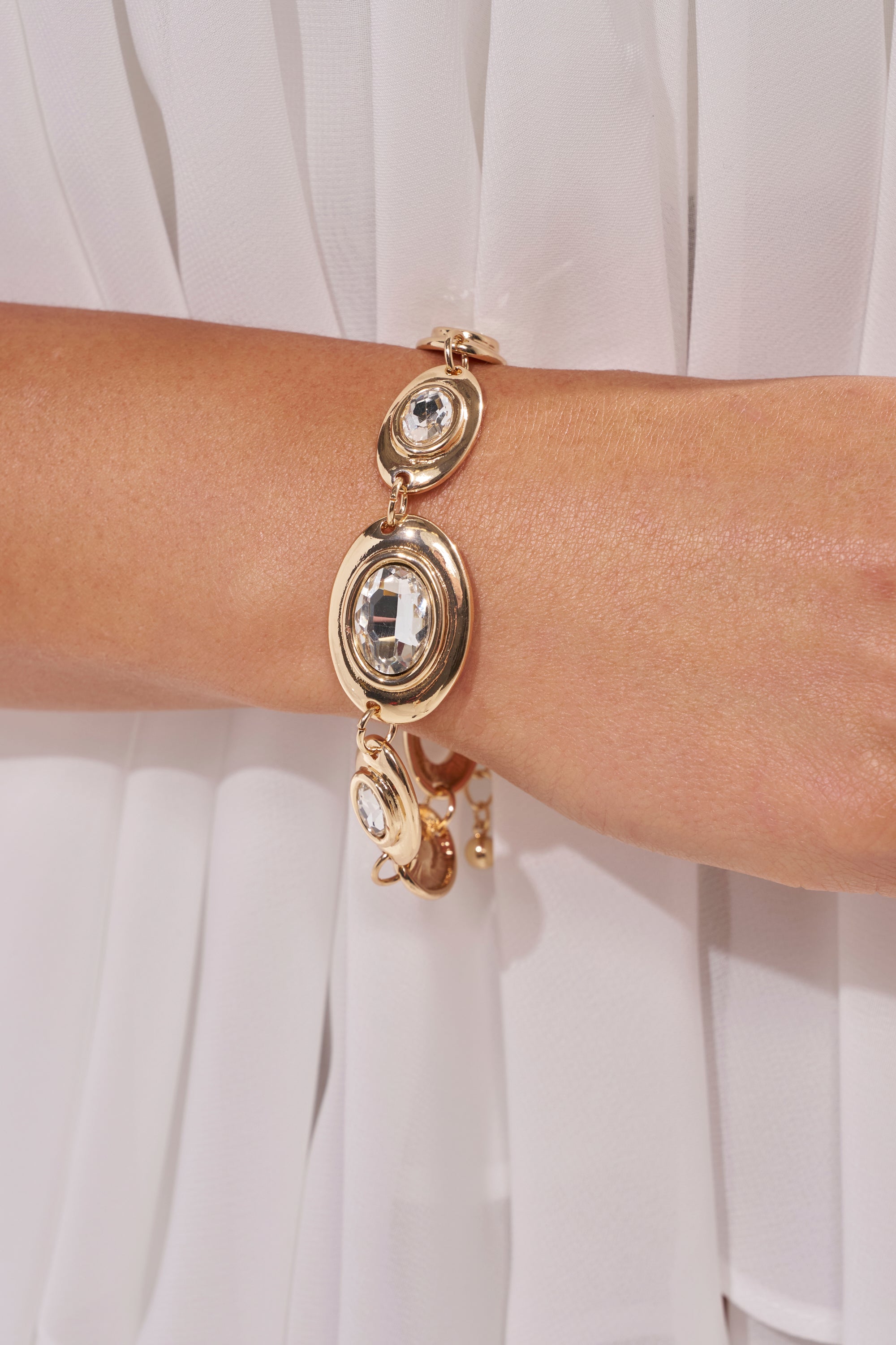 A close-up of the RUNWAY READY BRACELET IN GOLD, featuring large round clear gemstones, worn on a wrist over white pleated fabric. 1
