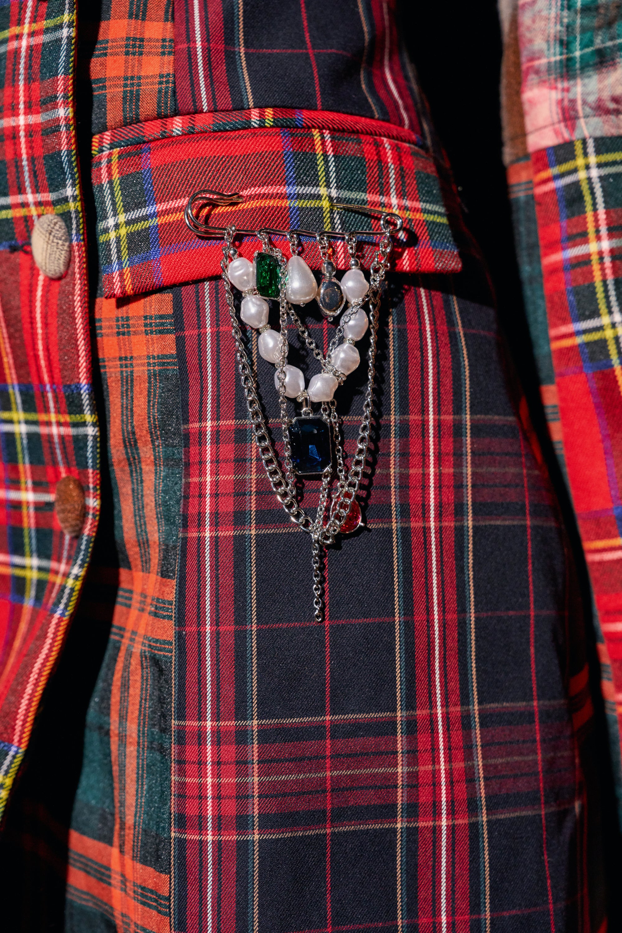 The SHES SO MOD PEARL BROOCH, a punk-inspired safety pin featuring layered silver chains, white pearls, and green and blue rectangular gemstones, is attached to a red and black plaid jacket.
