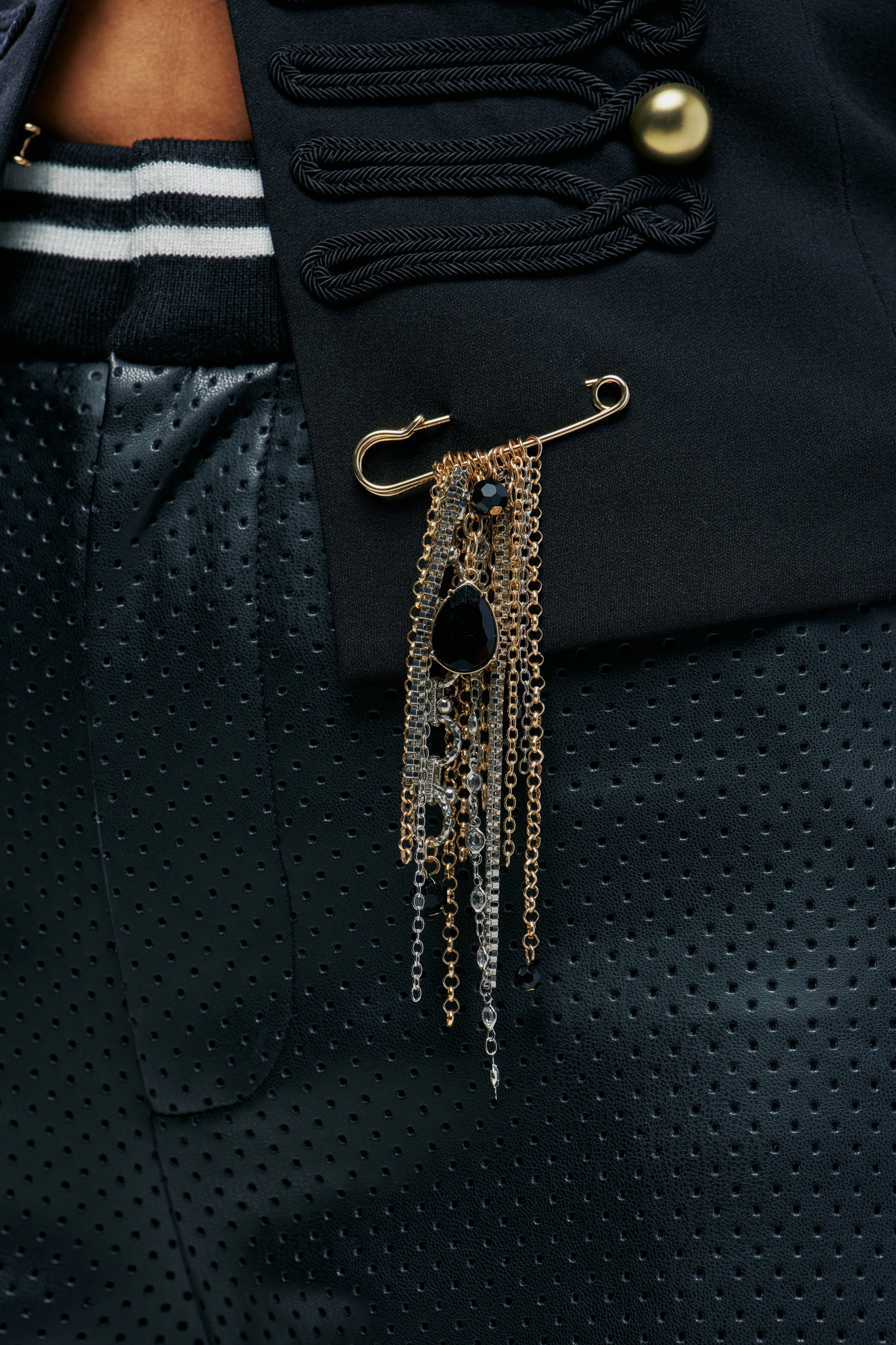 A MIXED MEDIA BROOCH featuring a maximalist gold safety pin with hanging gold and silver chains, accented by black teardrop and round beads, pinned to a black jacket.