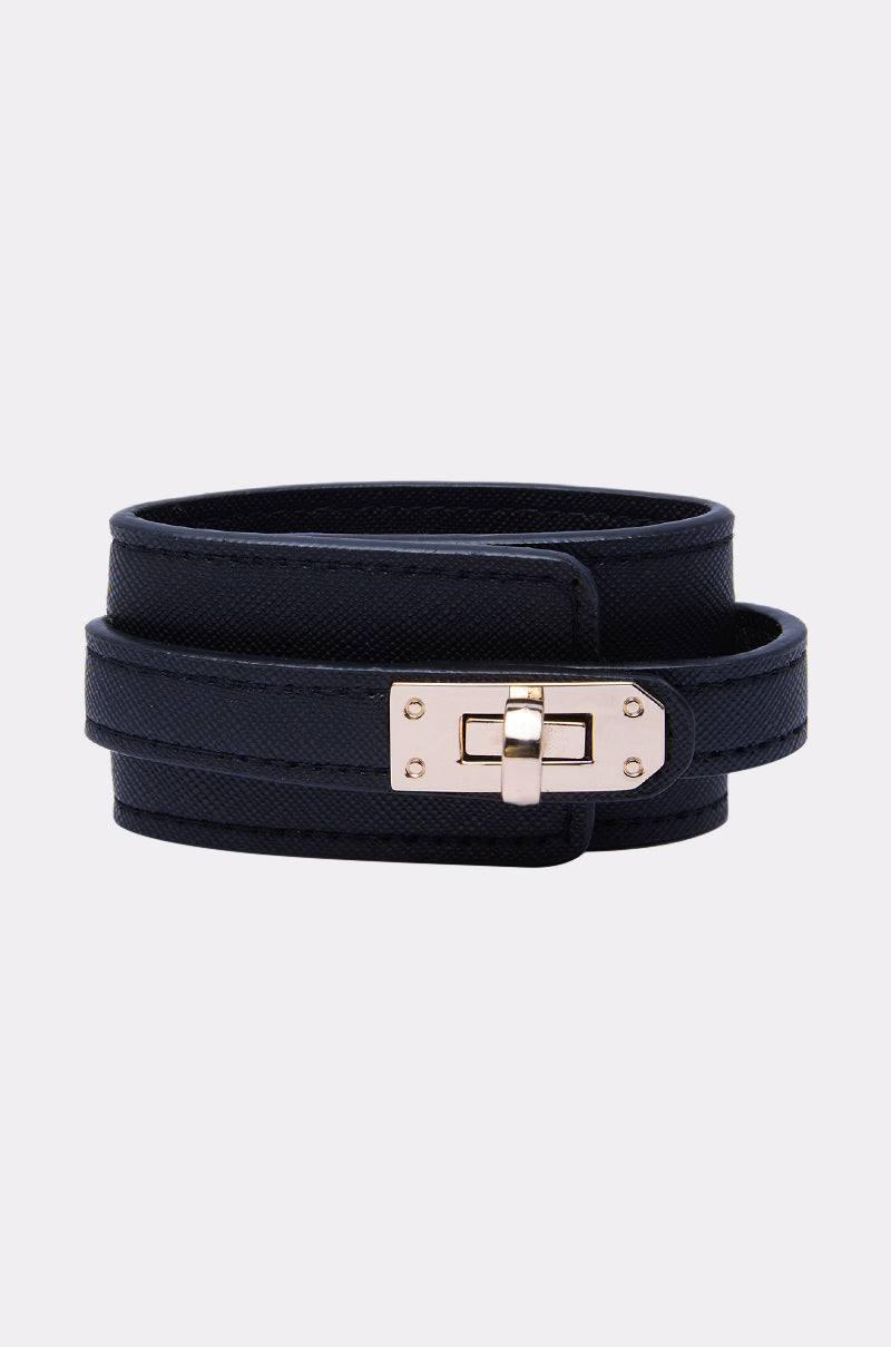 A DESIGNER BRACELET, featuring black textured leather and a gold rectangular buckle, is coiled neatly on a plain light gray background.