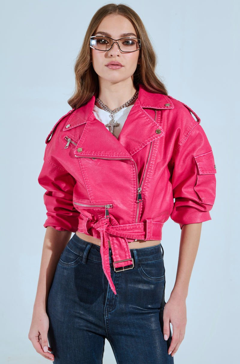 A long-haired person wearing glasses and the PINKY UP MOTO hot pink faux leather jacket with a detachable waist belt stands against a light blue background, accessorized with a silver necklace and dark jeans.