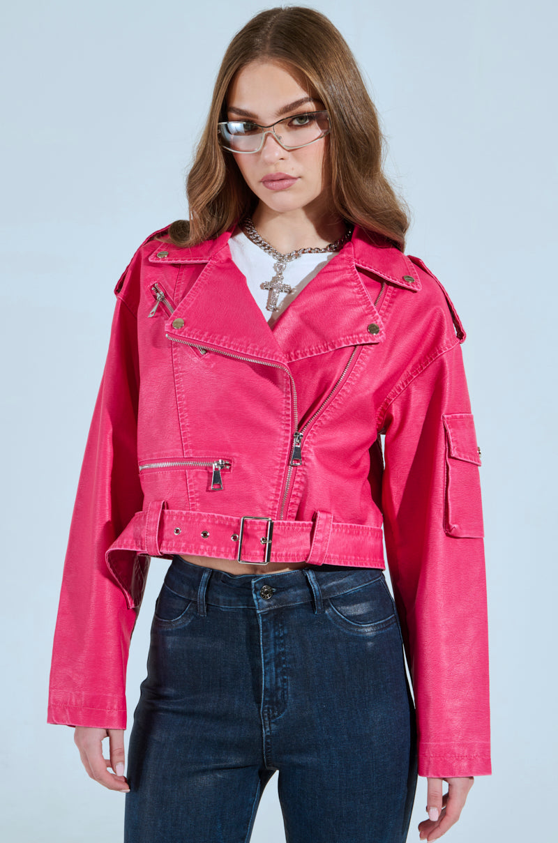 A woman with long brown hair wears sunglasses, a silver necklace, dark jeans, and the PINKY UP MOTO—a hot pink faux leather jacket with a cropped boxy silhouette and detachable waist belt—while standing against a plain background.