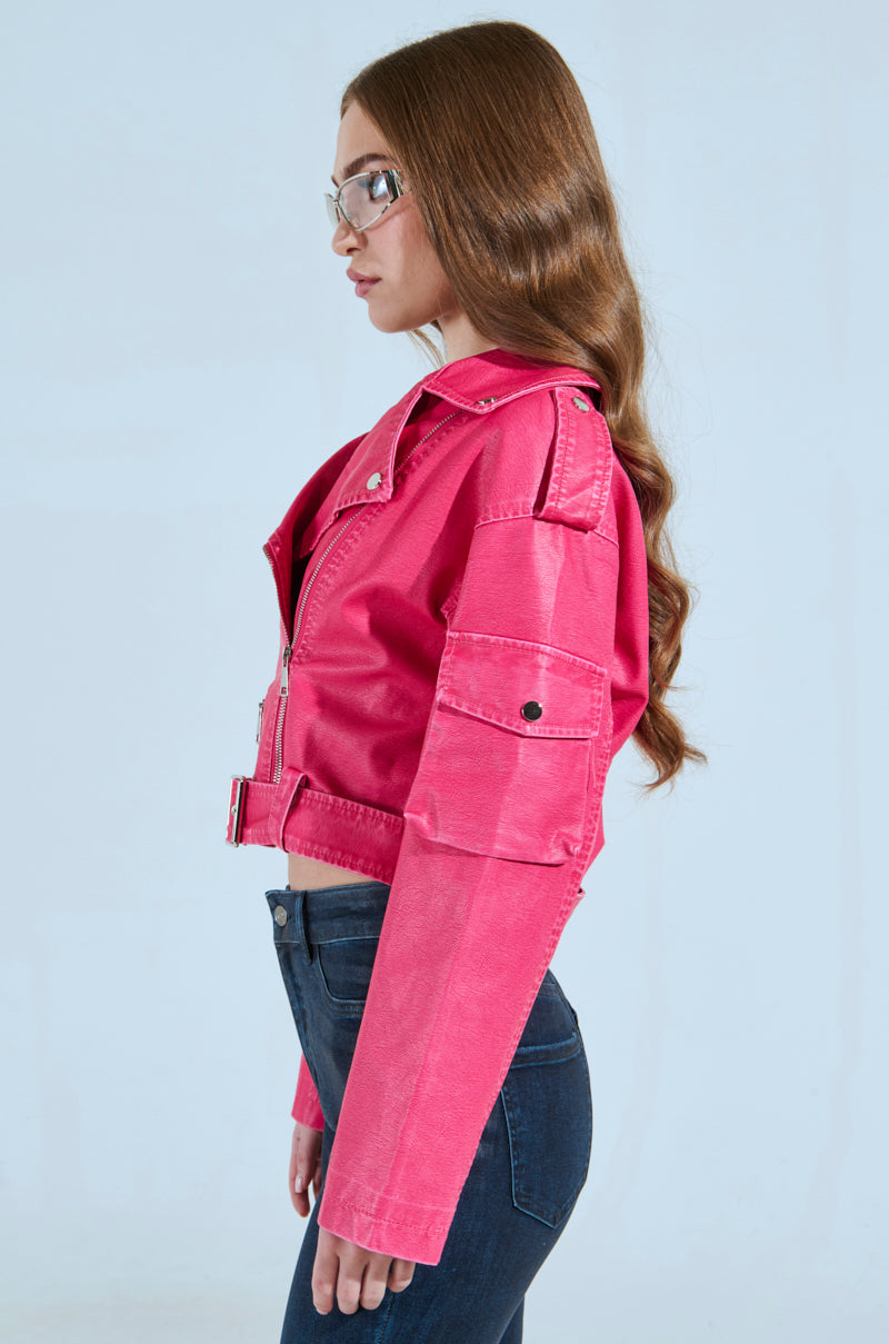 A woman with long brown hair, glasses, and dark blue jeans wears the PINKY UP MOTO, a hot pink faux leather jacket with a cropped boxy silhouette. She stands sideways against a light background, hands at her sides.
