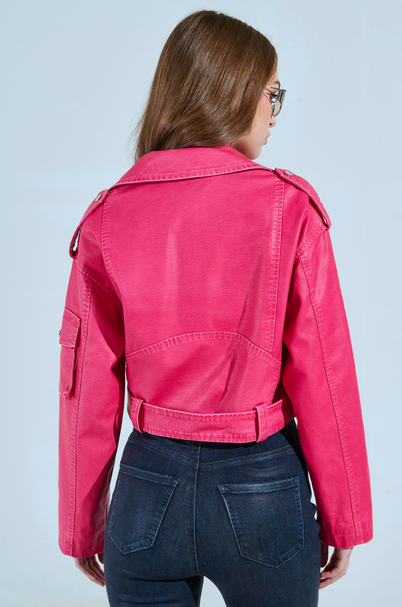 With long brown hair, a person faces away against a light blue background, wearing the PINKY UP MOTO hot pink faux leather jacket with a detachable waist belt, shoulder epaulets, and sleeve pocket, paired with dark blue jeans in a cropped boxy silhouette.
