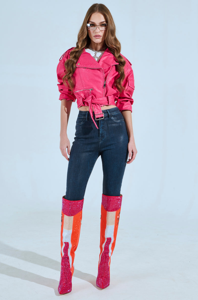 Wearing the PINKY UP MOTO hot pink faux leather jacket with a detachable waist belt, paired with dark skinny jeans and tall pink and orange boots, the person stands confidently against a light background. Their long hair and glasses complete this bold fashion statement.