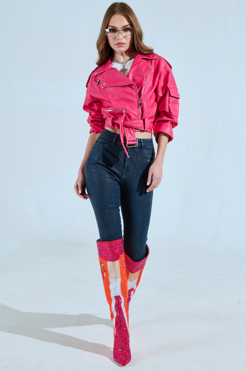 A woman confidently poses in the PINKY UP MOTO hot pink faux leather jacket with a cropped boxy silhouette over high-waisted dark jeans and tall, sparkly pink and orange boots. She finishes the look with sunglasses on her wavy brown hair, set against a plain white studio backdrop.
