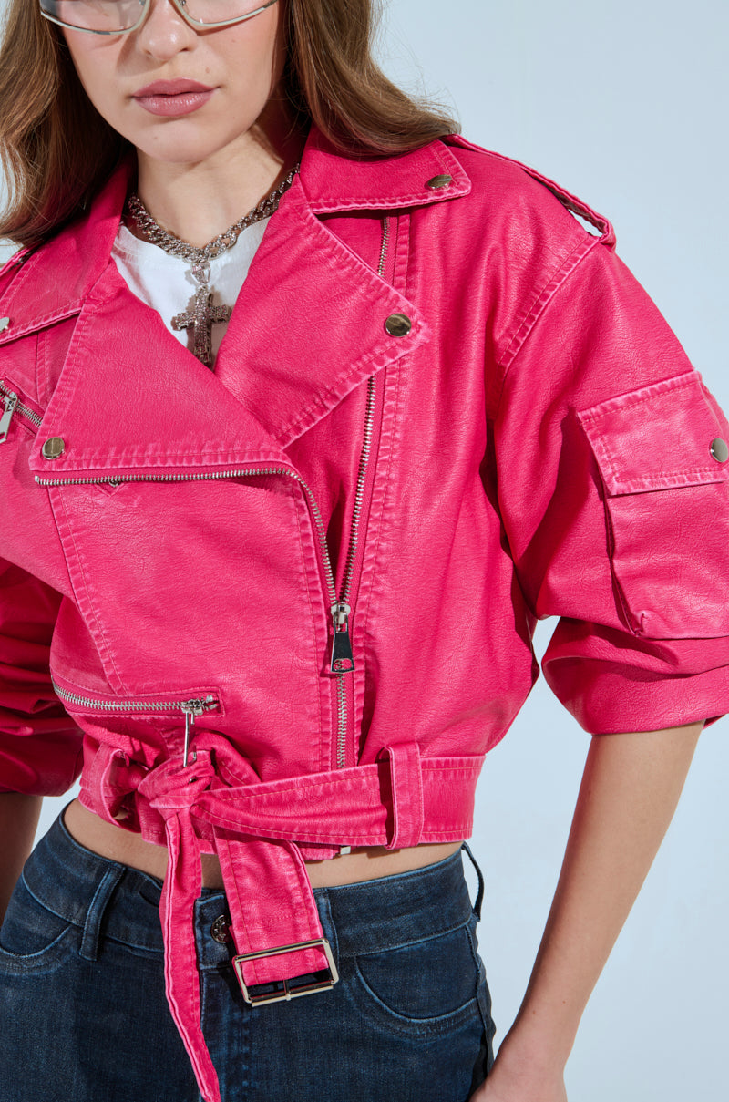 The individual with long hair and glasses is dressed in the PINKY UP MOTO hot pink faux leather jacket with detailed zippers, pockets, and a detachable waist belt over dark jeans. A silver necklace with a large cross pendant completes the look.