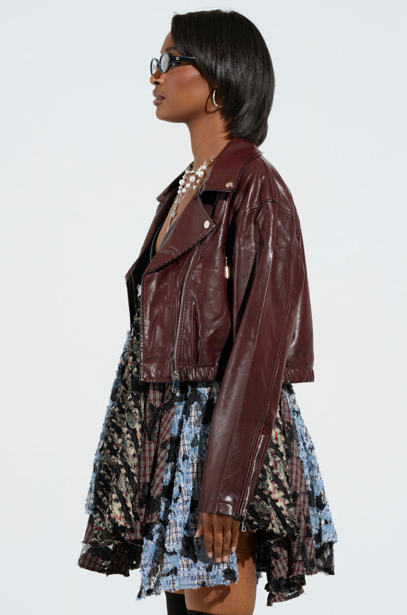 An individual with short dark hair and glasses is wearing the KALI MOTO faux leather jacket in maroon, enhanced with metallic accents, layered over a patterned dress showcasing a blue and black design. Their cropped silhouette is photographed from the side profile as they gaze to the right.