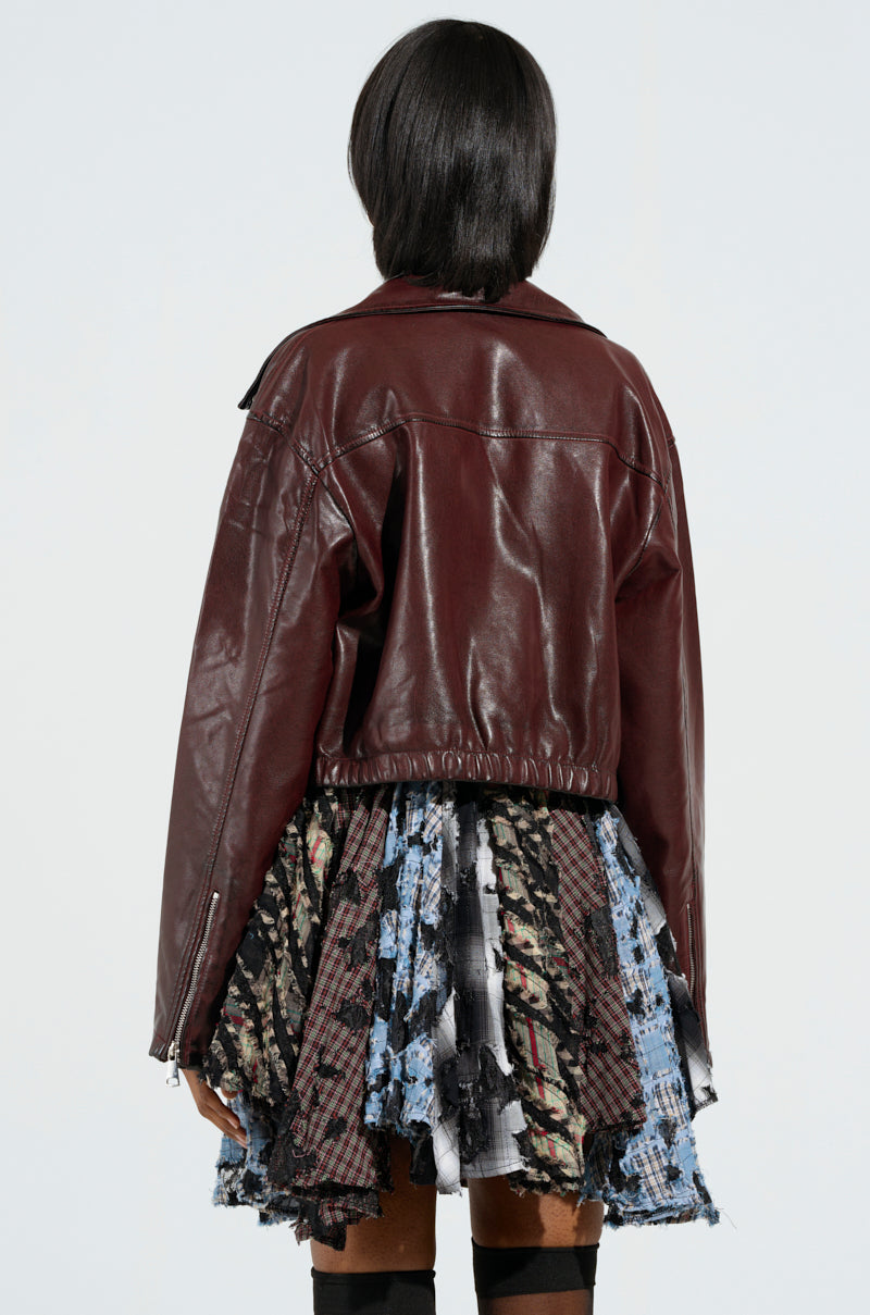 A person with short dark hair is wearing the KALI MOTO brown faux leather jacket, which features metallic hardware, over a multicolored patterned skirt. They are facing away, highlighting the cropped silhouette of their outfit against a plain background.