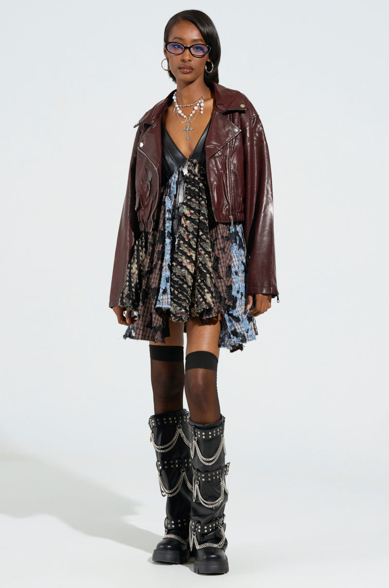 A person dressed in the KALI MOTO ensemble features a patterned dress, a maroon faux leather jacket enhanced with metallic hardware, and black knee-high boots embellished with chains. They accessorize with glasses and layered necklaces, posing against a plain background.