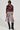 A person wearing the TEDDI MOTO jacket in a rich burgundy faux leather, showcasing its belted waist design, is teamed with a fringed skirt and black knee-high boots. They sport short hair and large sunglasses, exuding confidence as they stand against a plain background.