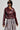 A person is striking a pose in the stylish TEDDI MOTO maroon faux leather jacket, showcasing its cropped silhouette with metal accents and long fringes, paired with oversized black sunglasses. The plain gray background accentuates the ensemble.