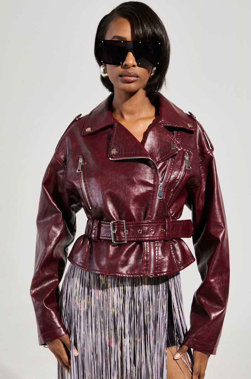 A woman wears the TEDDI MOTO, a shiny, deep red faux leather jacket featuring a belted waist, elegantly paired with a fringed skirt. Her short black hair enhances the look of her large, dark sunglasses against a plain light-colored background.