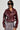 A woman wears the TEDDI MOTO, a shiny, deep red faux leather jacket featuring a belted waist, elegantly paired with a fringed skirt. Her short black hair enhances the look of her large, dark sunglasses against a plain light-colored background.
