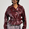 A woman wears the TEDDI MOTO, a shiny, deep red faux leather jacket featuring a belted waist, elegantly paired with a fringed skirt. Her short black hair enhances the look of her large, dark sunglasses against a plain light-colored background.