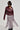 Wearing the TEDDI MOTO jacket, a person exudes confidence with its burgundy faux leather finish, belted waist, and long fringes. Sporting short hair and large, dark sunglasses, they complement the look with tall boots while striking a pose against a plain background.