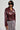 A woman wearing the TEDDI MOTO burgundy faux leather jacket and large black sunglasses stands against a plain background. The jacket's cropped silhouette, featuring a belt and silver zipper details, complements her fringed skirt. Her expression is neutral.