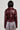A person with black hair is seen from behind sporting the TEDDI MOTO, a shiny, dark red faux leather jacket, alongside a skirt adorned with fringe details and a multicolored pattern. The scene highlights a cropped silhouette against a plain light gray backdrop.