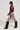 A person wearing the TEDDI MOTO burgundy faux leather moto jacket, a silver fringe skirt, large sunglasses, and tall black boots walks confidently. The cropped silhouette of the jacket adds a bold touch to their look. The background is plain and well-lit, casting a shadow on the floor.