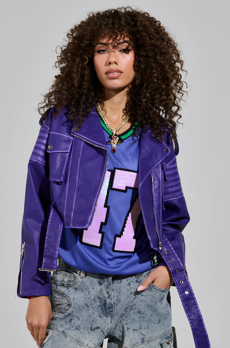 A person with curly hair is wearing a LEILANI MOTO faux leather jacket over a sports jersey featuring the number 17. They complement their look with layered necklaces and acid-wash jeans, standing against a plain backdrop.