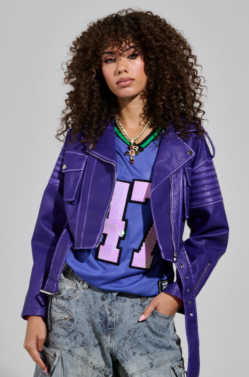 A person with curly hair confidently wearing the LEILANI MOTO, a purple faux leather jacket with an acid wash trim, over a blue sports jersey featuring the number 47. They sport layered necklaces and baggy jeans against a plain background.