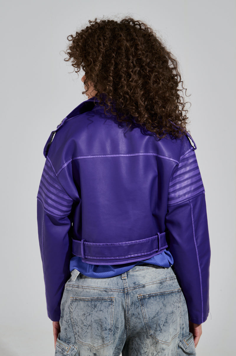 A person with curly hair is seen from behind, wearing the striking LEILANI MOTO jacket in vibrant purple faux leather. This jacket features shoulder straps and horizontal stitching on the arms, and its cropped silhouette pairs perfectly with acid wash trim jeans, all set against a neutral gray background.