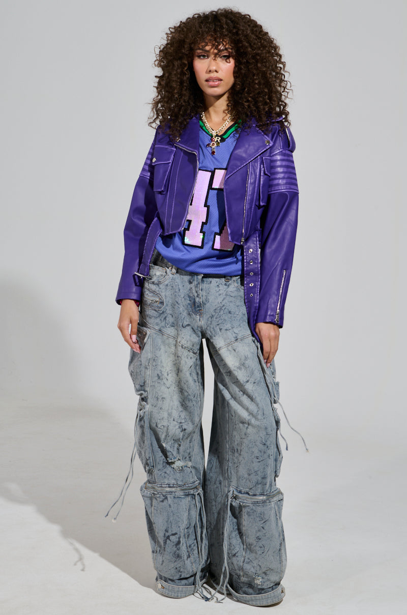 Confidently posed against a plain background, a person with curly hair wears the LEILANI MOTO, a purple faux leather jacket, over a blue sports jersey featuring the number 41, complemented by oversized acid-wash trim denim cargo pants. They complete their look with layered necklaces.