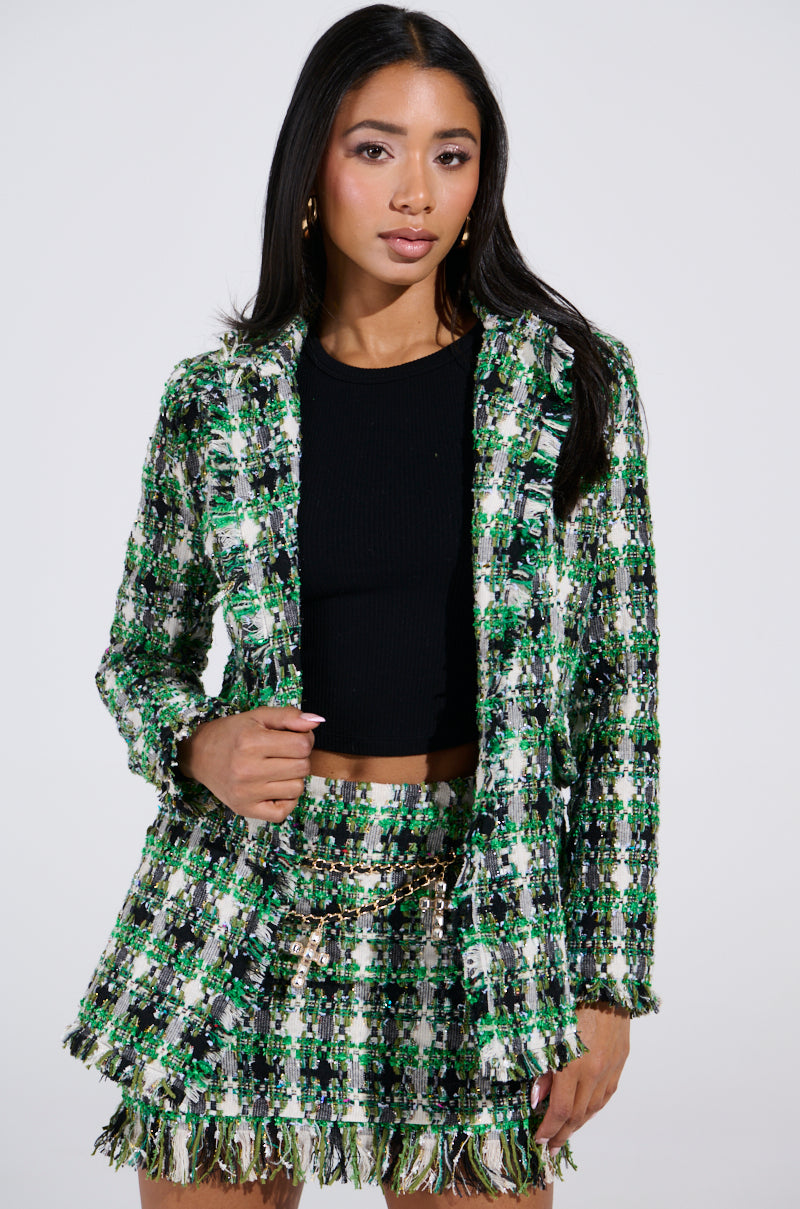 A person confidently poses against a plain background, showcasing the GEORGIA TWEED BLAZER in green, black, and white tweed, paired with a matching skirt and black top. Their long black hair perfectly complements the ensemble.
