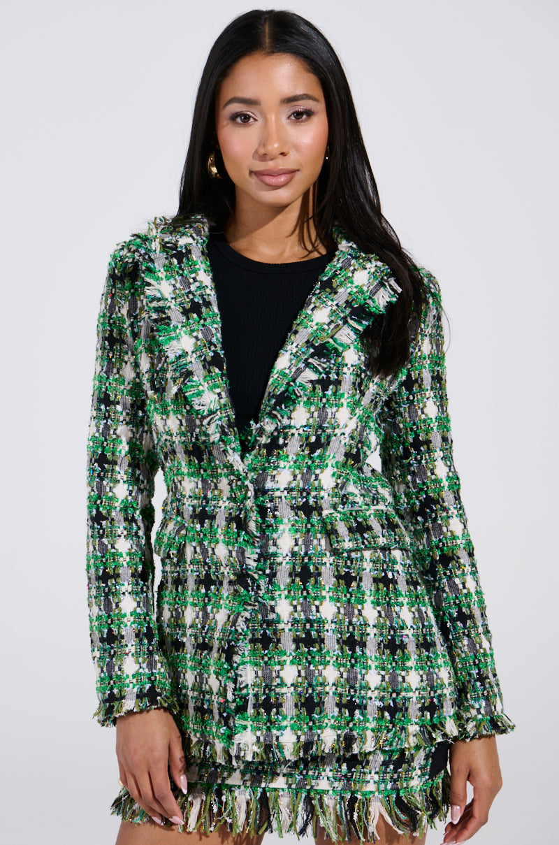 A person with long dark hair wears a green and white plaid AKIRA Label jacket with fringe over a black top, resembling the GEORGIA TWEED BLAZER style, standing elegantly against a plain white background.