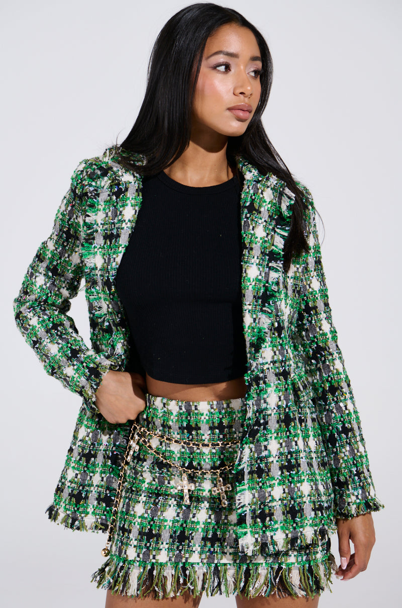 Dressed in an AKIRA Label GEORGIA TWEED BLAZER with a matching skirt featuring gold chains, a person with long dark hair poses against a light background, hand on hip.