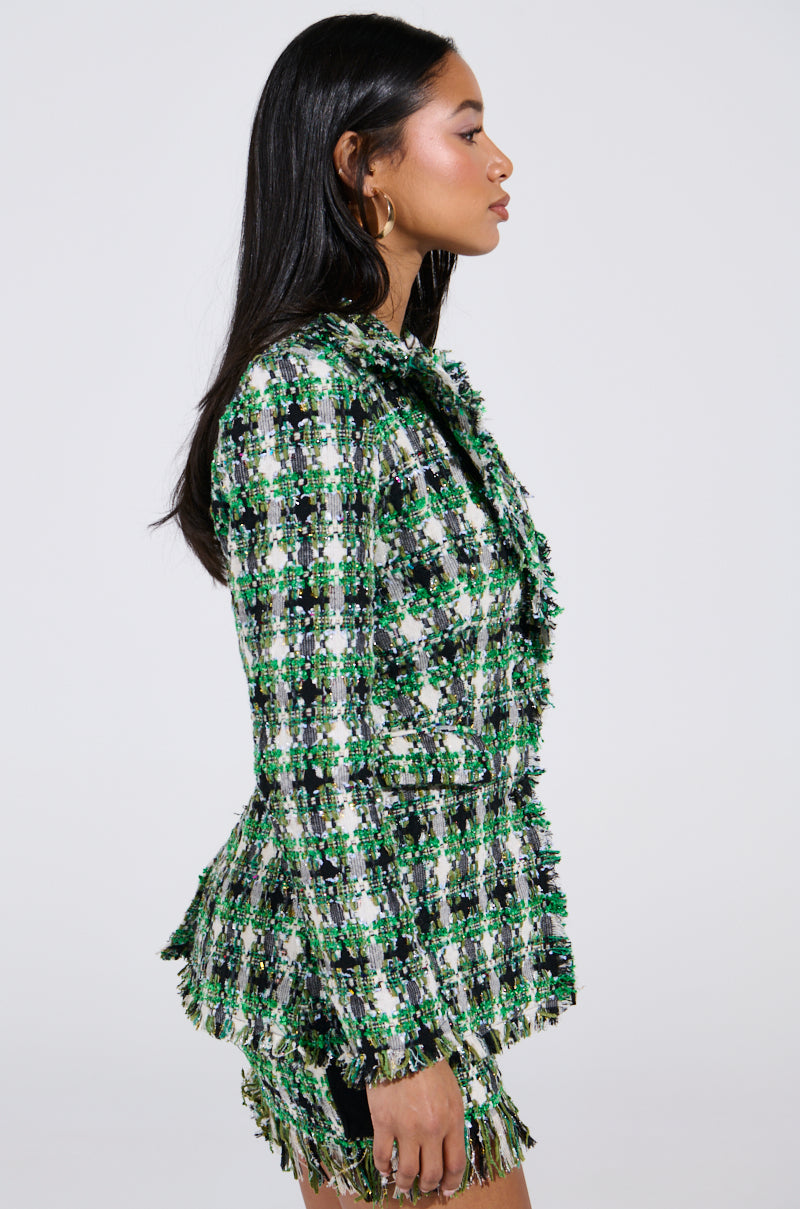 A woman with long dark hair in profile models the GEORGIA TWEED BLAZER, featuring a green and white checkered pattern with fringe details. She accessorizes with gold hoop earrings, against a plain white backdrop.