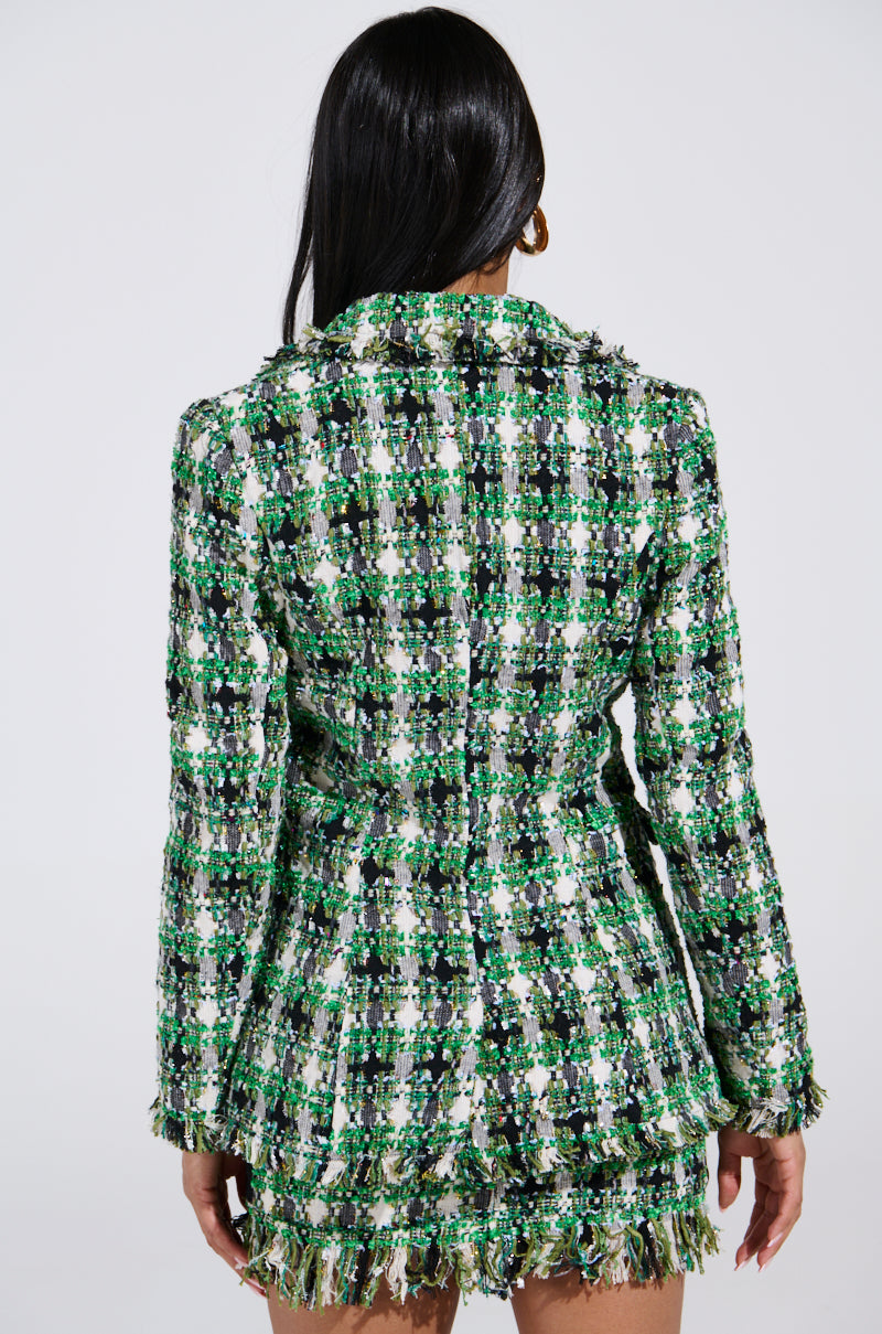 A person shows off an AKIRA Label GEORGIA TWEED BLAZER in green and white, with a textured pattern and fringed edges. Their long, dark hair contrasts against the plain, light-colored background.