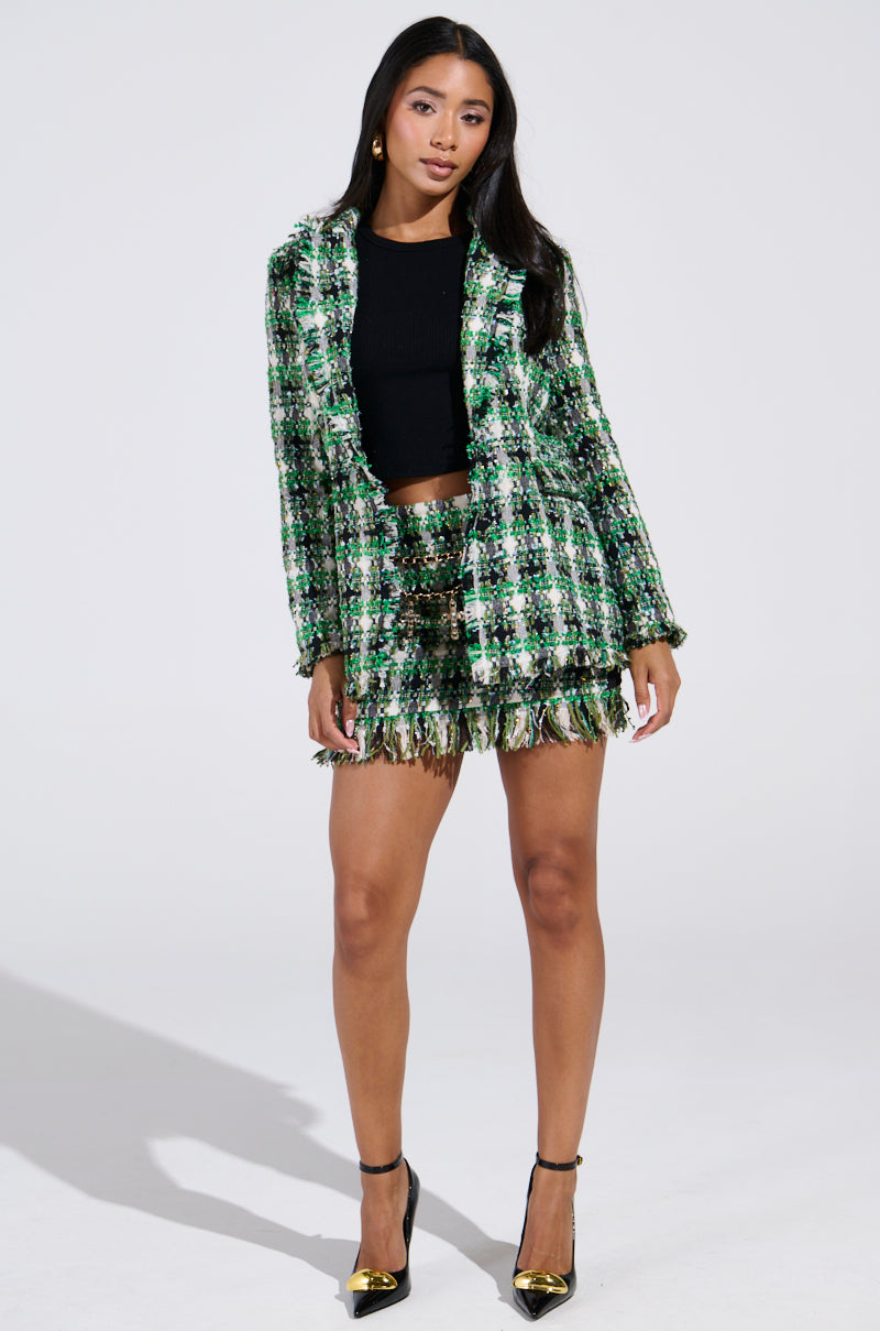 A woman exudes confidence against a plain backdrop, wearing the GEORGIA TWEED BLAZER set in green and black plaid tweed with fringe. She pairs the ensemble with a black top, and black heels with gold tips, her long hair cascading down stylishly.