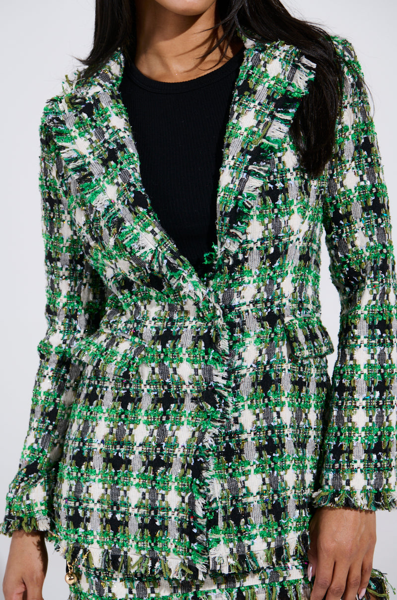 The woman flaunts a striking AKIRA Label GEORGIA TWEED BLAZER, adorned with a green, black, and white plaid pattern. This chic piece features a frayed hem and fitted cut over her black top.