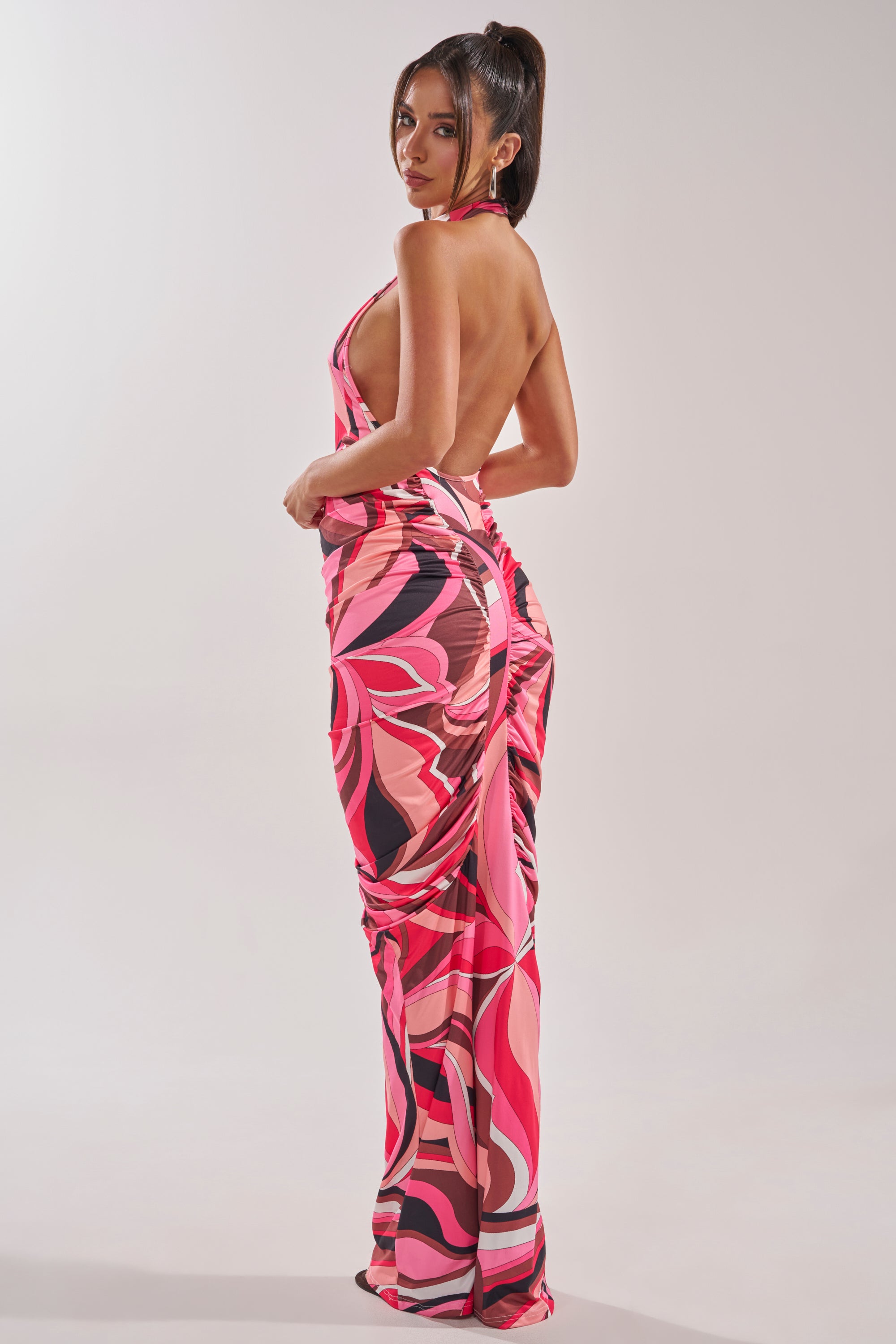 A woman stands sideways against a plain background, wearing the PERFECTLY EXTRA HIGH NECK PRINTED MAXI DRESS with a backless, halter-neck design and bold pink, red, and black abstract swirl patterns. 1