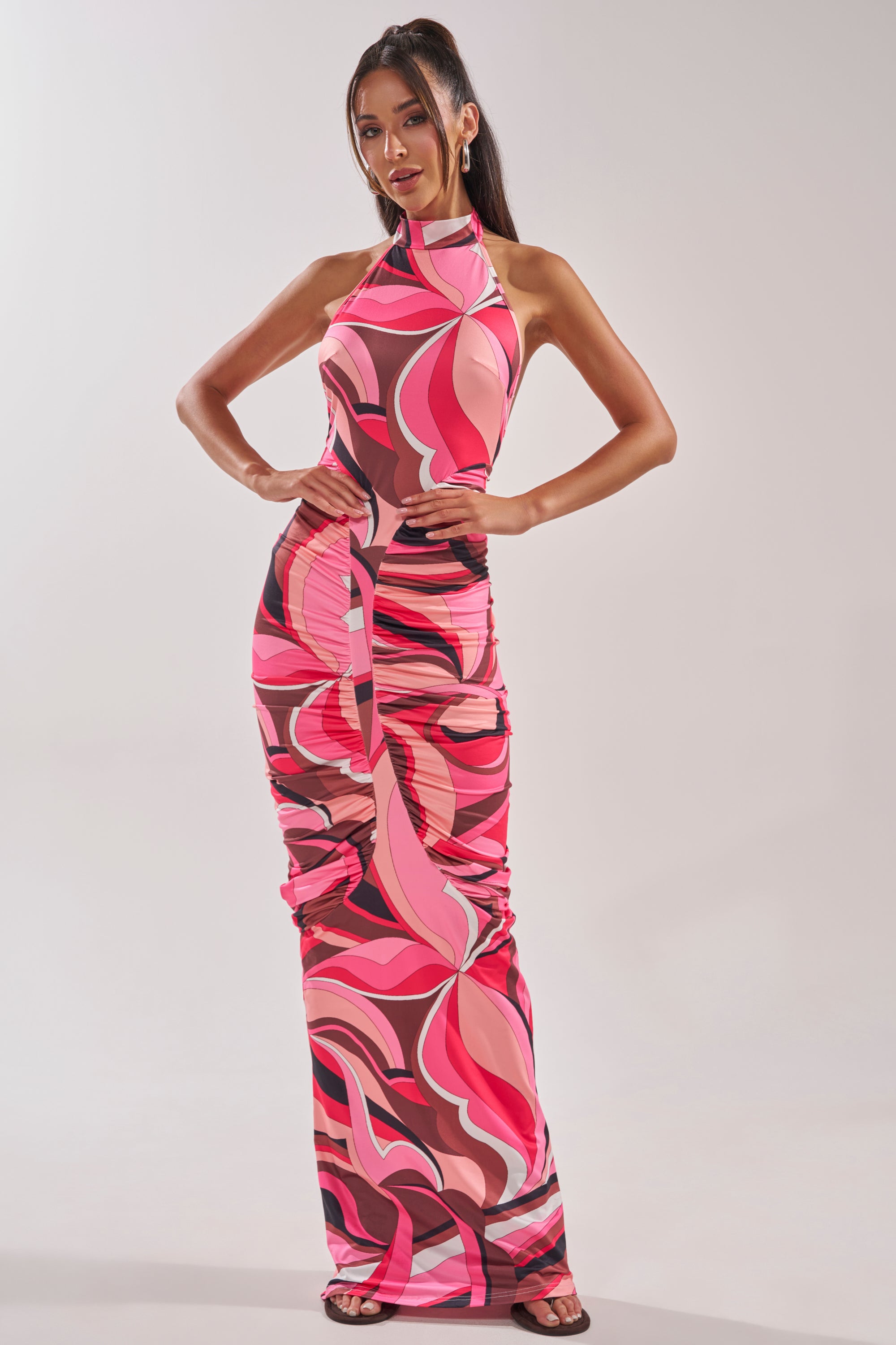 Woman poses in the PERFECTLY EXTRA HIGH NECK PRINTED MAXI DRESS, a sleeveless, form-fitting maxi with bold pink, red, and black abstract swirls. She pairs it with dark sandals and a high ponytail for a striking retro glam look. 2