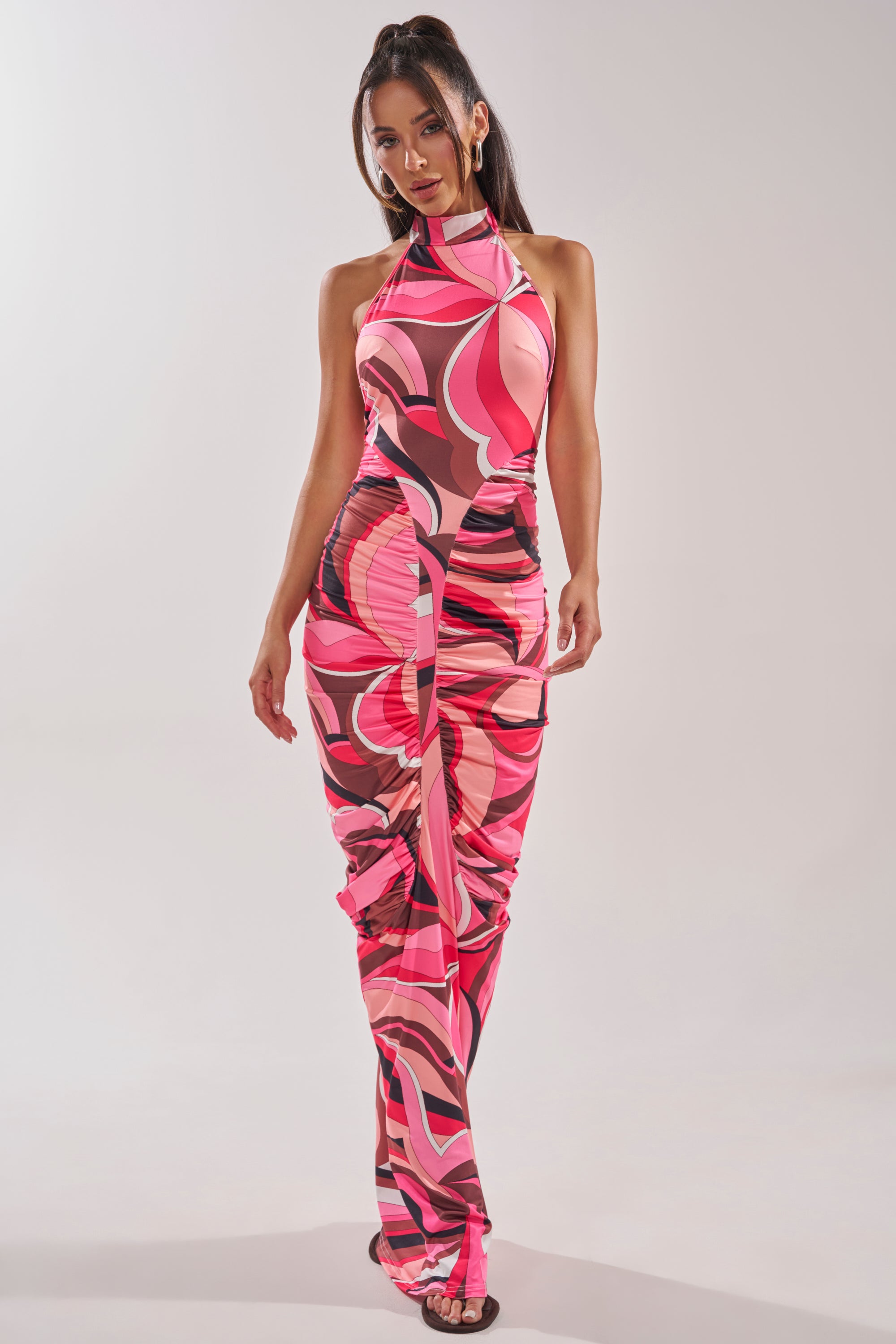 A woman wears the PERFECTLY EXTRA HIGH NECK PRINTED MAXI DRESS, featuring a bold pink, black, and white abstract print. Her long dark hair is styled half-up, and she completes the look with chic black sandals. 3