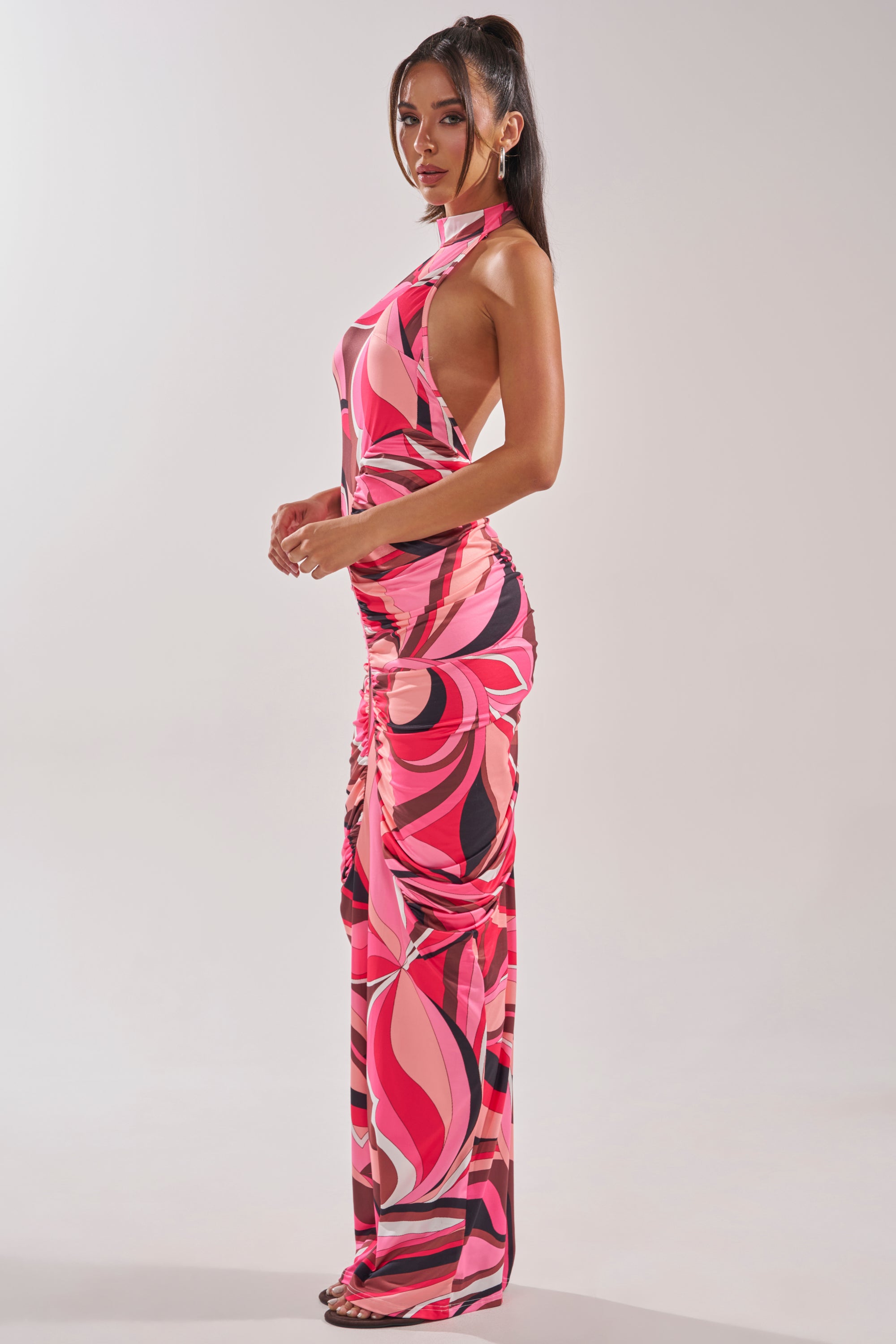 A woman with long dark hair in a high ponytail poses sideways against a plain light background, wearing the PERFECTLY EXTRA HIGH NECK PRINTED MAXI DRESS featuring a sleeveless, form-fitting silhouette with bold pink, red, and black swirl patterns. 4