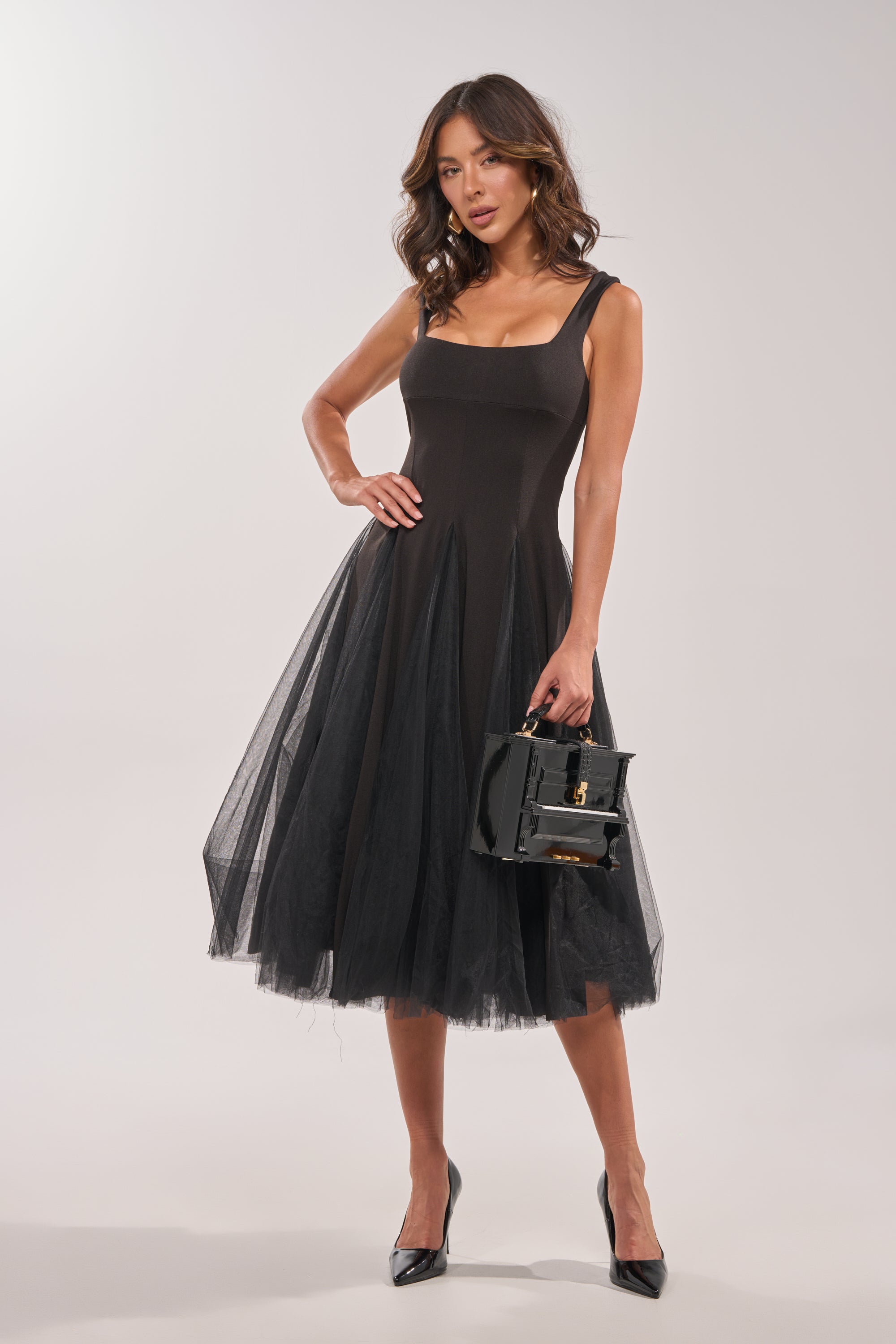 A woman wears the KNOW YOUR WORTH TULLE MIDI DRESS IN BLACK, featuring a sleeveless design and layered tulle skirt. She poses with one hand on her hip, holding a black handbag, black heels, and wavy shoulder-length hair against a white background.