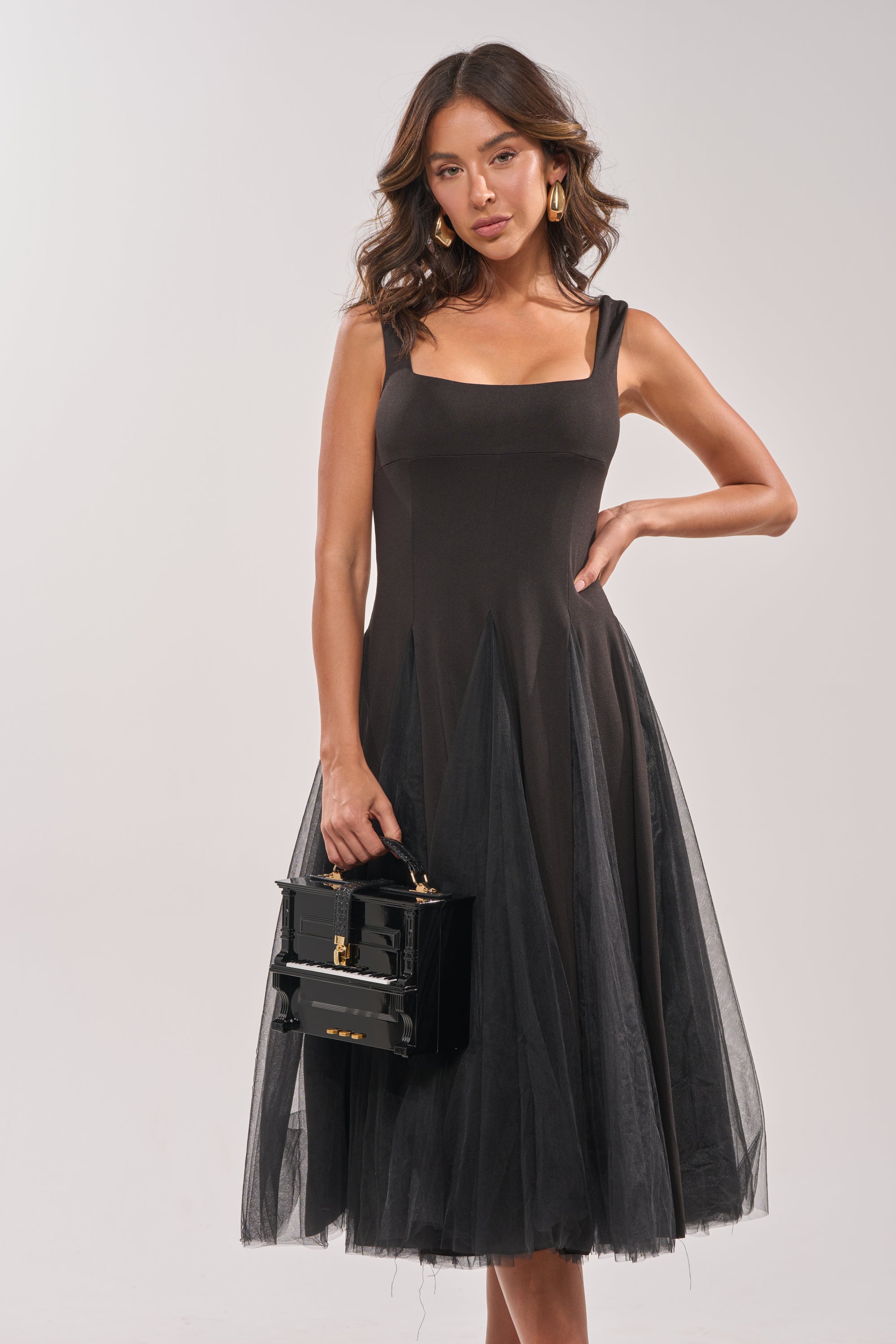 A woman wears the KNOW YOUR WORTH TULLE MIDI DRESS IN BLACK, featuring a sleeveless design and layered tulle skirt, accessorized with a small black handbag and gold earrings against a plain background.