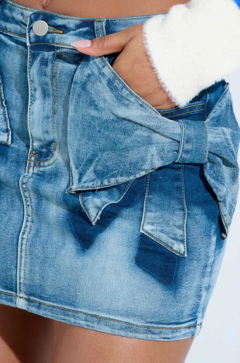 A person wears the PRETTY GIRL PROBLEMS MINI SKIRT, a blue faded denim mini skirt with a large bow applique. Paired with a white fluffy sweater, their hand casually rests in the pocket. The metallic button of the skirt gleams visibly.