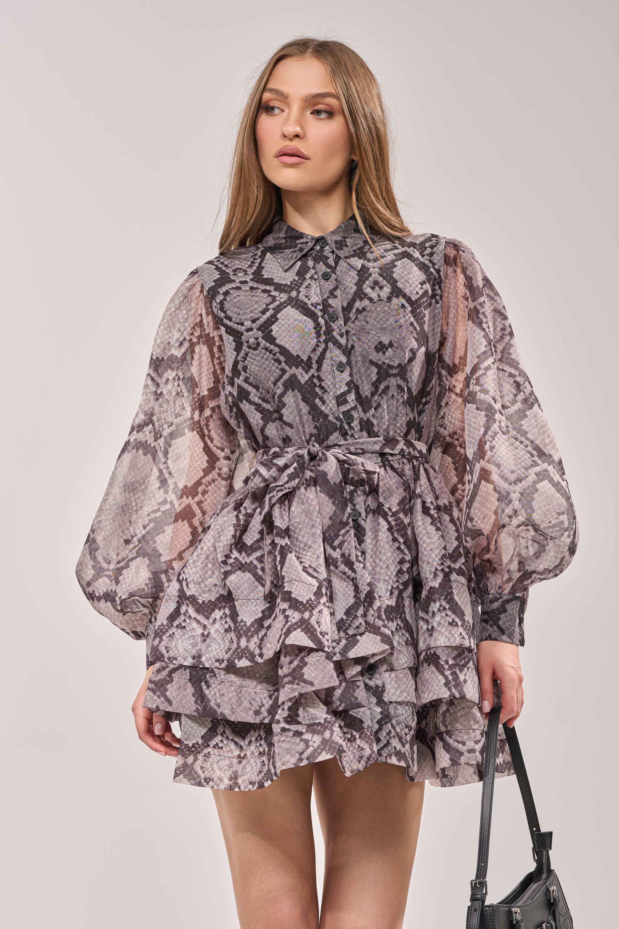 A woman with long hair wears the IT GIRL CHIFFON RUFFLE MINI DRESS IN SNAKE, featuring voluminous sleeves and a tie waist, holding a black handbag against a plain light background. 1