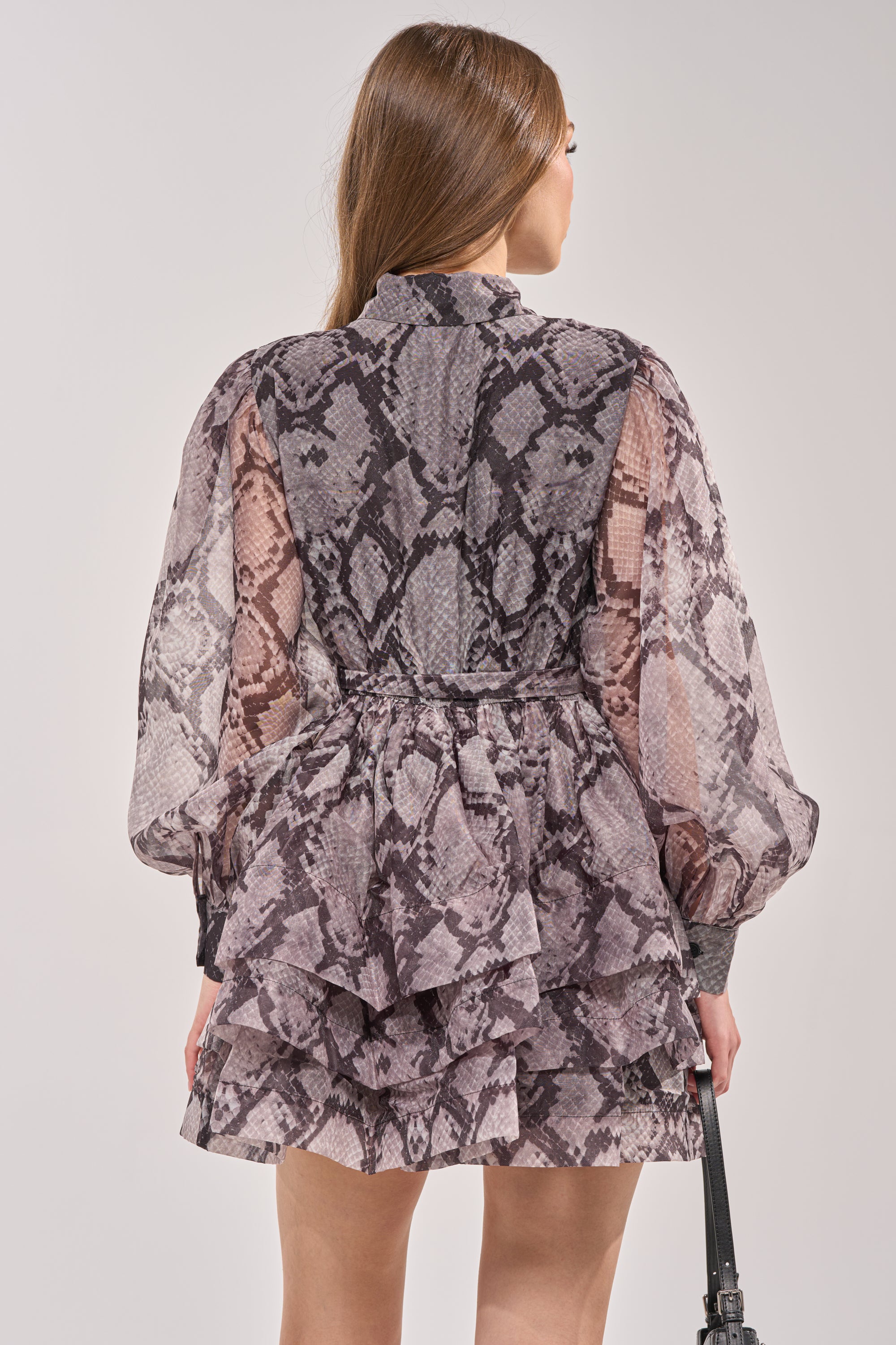 A woman with long brown hair is seen from the back, wearing the IT GIRL CHIFFON RUFFLE MINI DRESS IN SNAKE—a sheer, long-sleeved snake print mini dress with ruffles—paired with a black handbag. 3