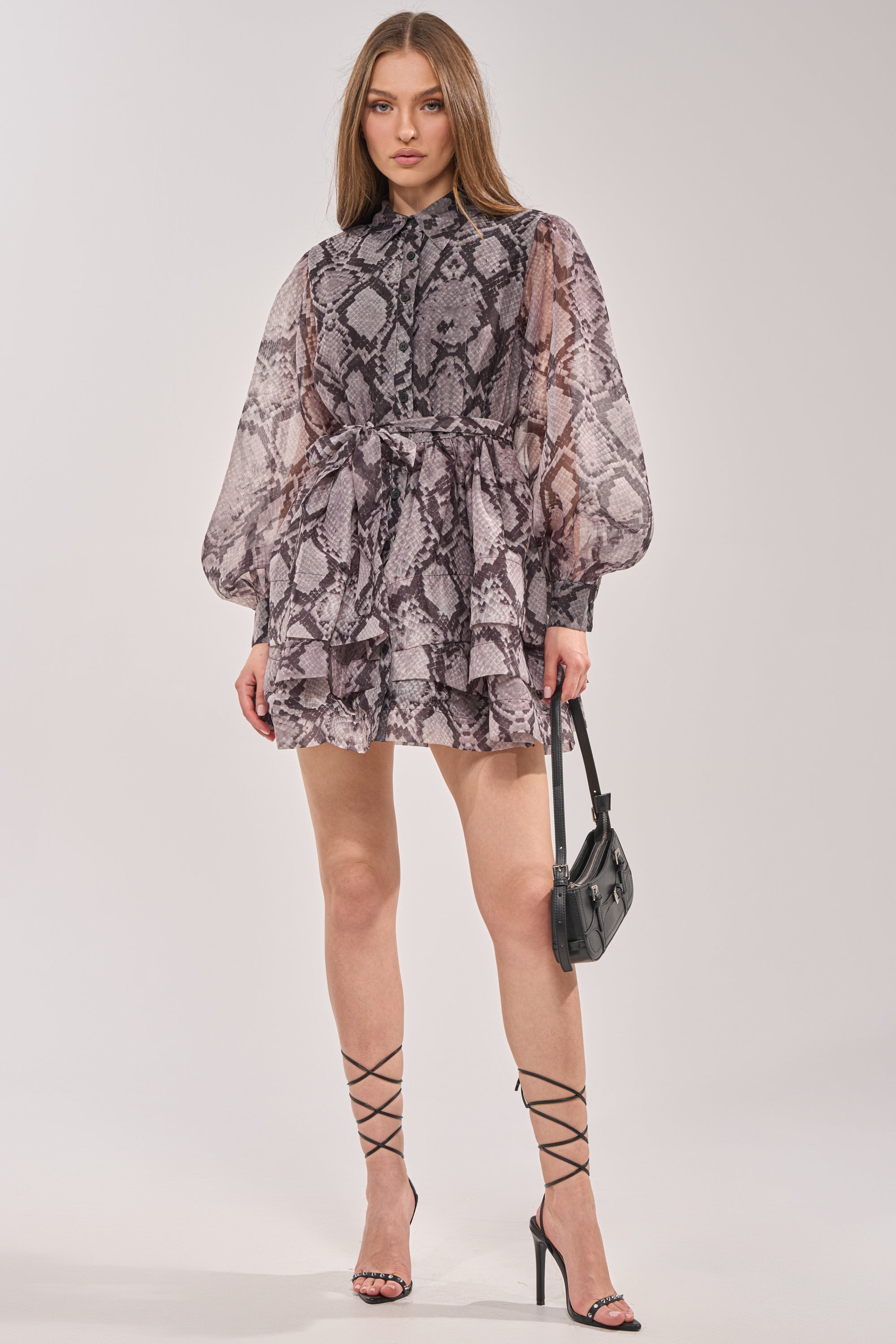 A woman wears the IT GIRL CHIFFON RUFFLE MINI DRESS IN SNAKE, featuring sheer long sleeves and a tiered skirt, paired with black strappy heels and a small black handbag, standing against a plain light background. 4