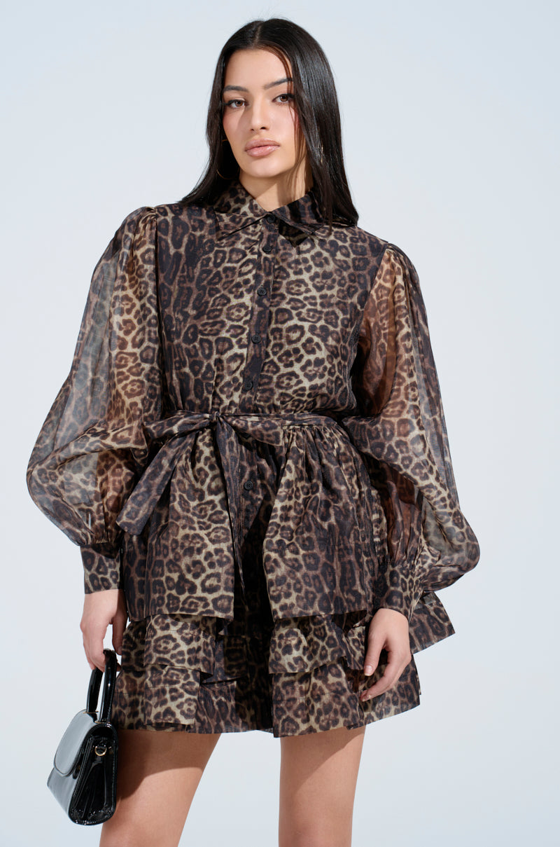 A person with long dark hair confidently wears the CHICEST OF CHIC leopard print mini dress, featuring sheer fabric, bishop sleeves, and a waist tie. They hold a small black handbag against a plain light background.