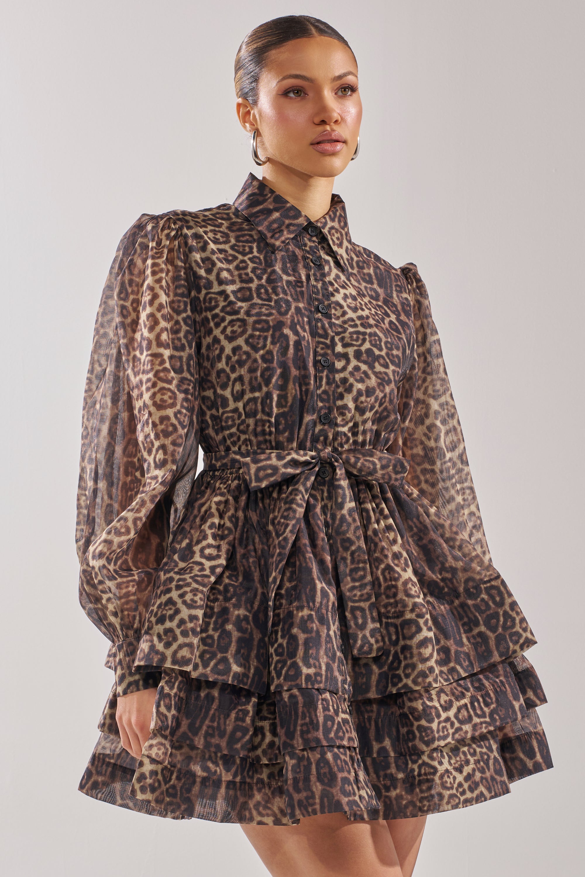 A woman wears the CHICEST OF CHIC LEOPARD PRINT MINI DRESS featuring bishop sleeves, a button-up front, and a tied waist belt, standing against a plain light background.