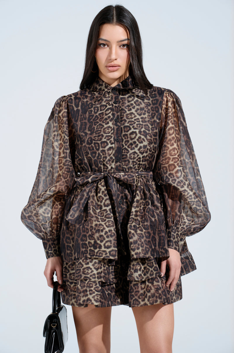 With long dark hair, a person wears the CHICEST OF CHIC LEOPARD PRINT MINI DRESS featuring bishop sleeves and a tied waist, holding a black handbag against a plain light blue background.