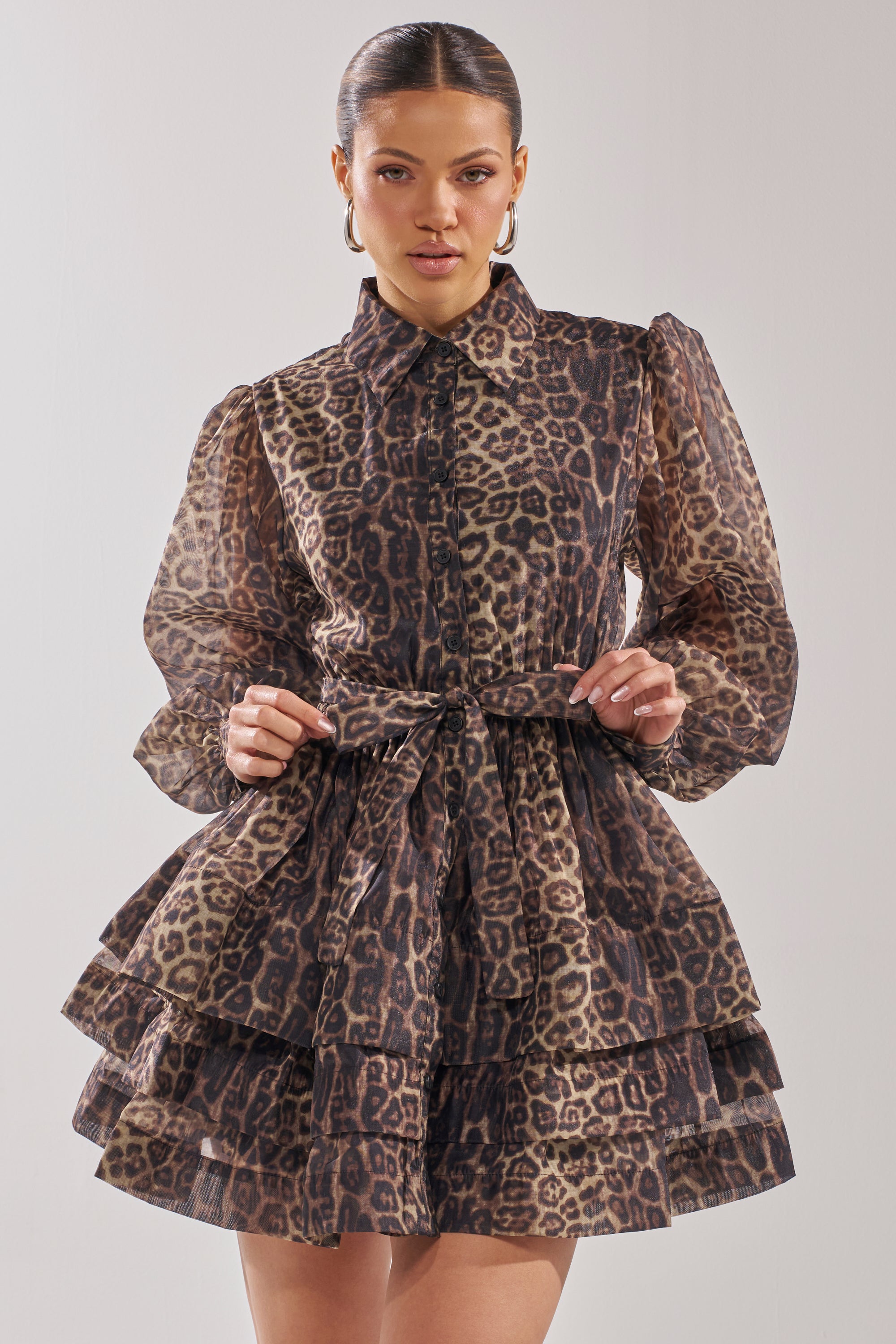 A woman models the CHICEST OF CHIC LEOPARD PRINT MINI DRESS, featuring bishop sleeves, a collared neckline, tied waist, and tiered skater silhouette against a plain light background.
