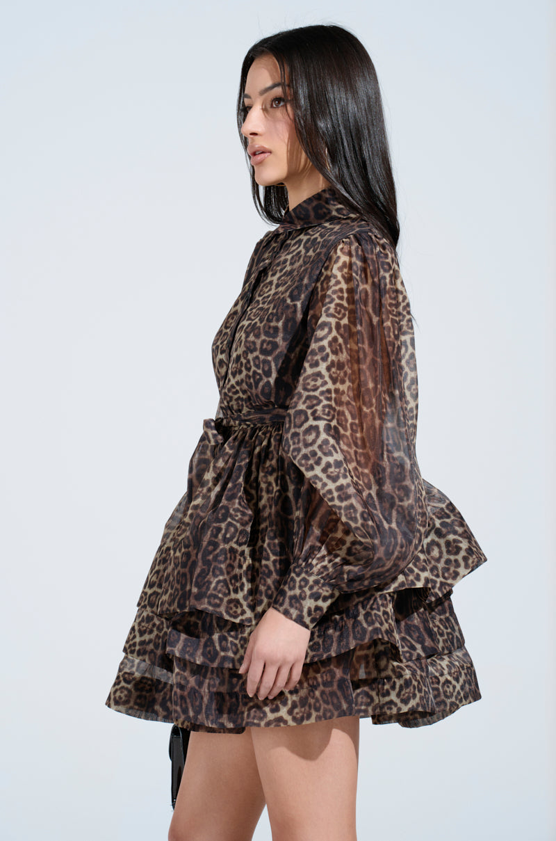 A woman with long dark hair models the CHICEST OF CHIC LEOPARD PRINT MINI DRESS. It features bishop sleeves, a high neckline, and bold style, all set against a plain light backdrop as she looks to the side.
